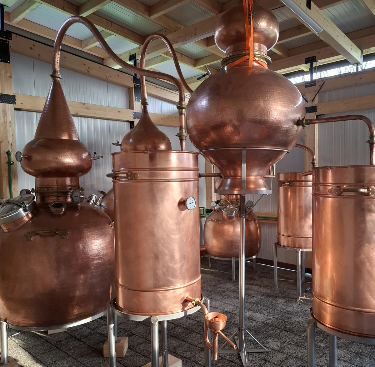 miedziane alembiki i aparatura do destylacji ustawione w drewnianej hali, skupione na detalach technicznych oraz zakrzywionych rurach łączących poszczególne zbiorniki