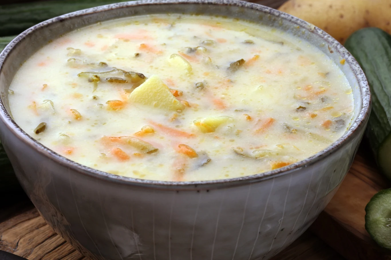 Miska kremowej zupy ogórkowej z kawałkami ziemniaków, tartą marchewką i widocznymi paskami ogórka kiszonego, całość o apetycznej, lekko zielonkawej barwie.