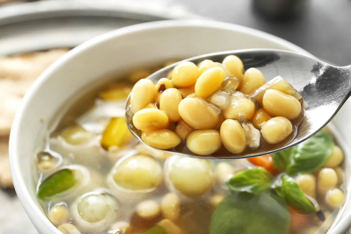 łyżka z ugotowaną białą fasolą trzymana nad miską pełną klarownej zupy z widocznymi kawałkami warzyw i listkiem bazylii