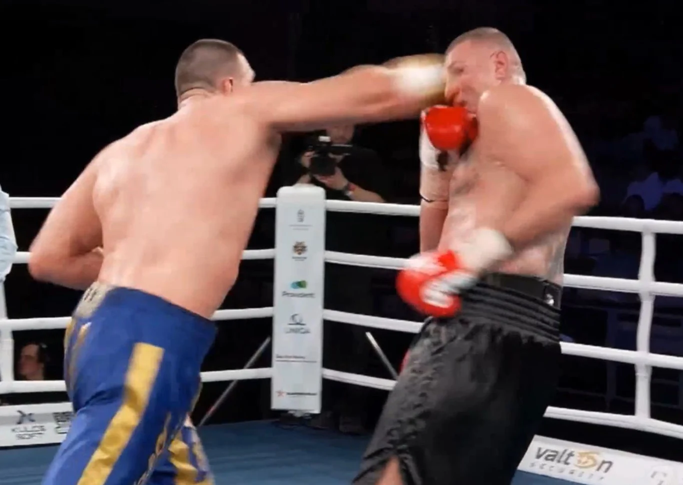 Dwóch bokserów walczy w ringu, jeden z nich zadaje mocny cios prosty w twarz przeciwnika, na tle lin ringu i aparatu fotograficznego skierowanego na akcję.