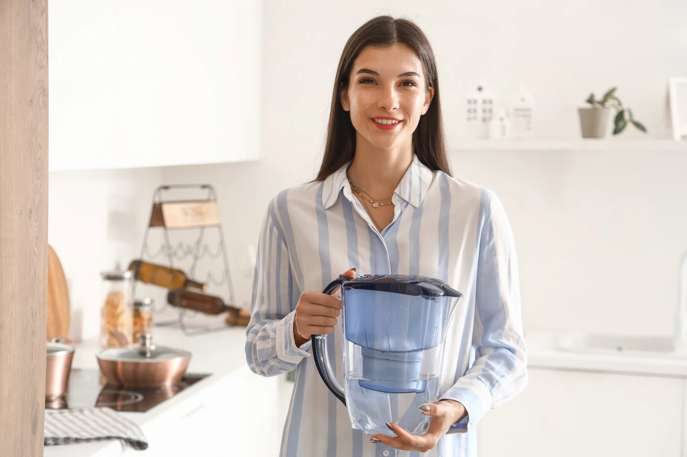 Dzbanek filtrujący powinniśmy regularnie czyścić. Pewien domowy sposób pomoże nam usunąć z niego osad i kamień. Młoda kobieta w jasnej koszuli w paski trzyma w rękach dzbanek filtrujący wodę. Stoi w jasnej kuchni. Czyszczenie dzbanka.