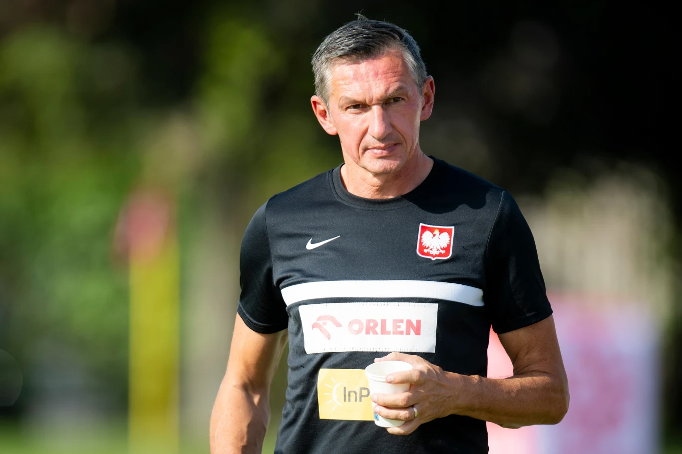 mężczyzna w czarnej sportowej koszulce z godłem Polski oraz logo Orlen, trzymający kubek w jednej ręce, stoi na zewnątrz na tle rozmytego zielonego otoczenia