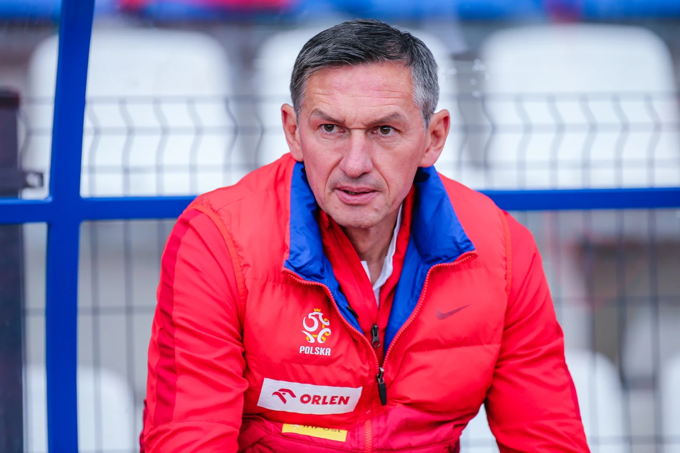 mężczyzna w czerwonej kurtce z logo reprezentacji Polski oraz napisem Orlen siedzi na ławce na tle stadionowych krzesełek, patrząc z poważnym wyrazem twarzy