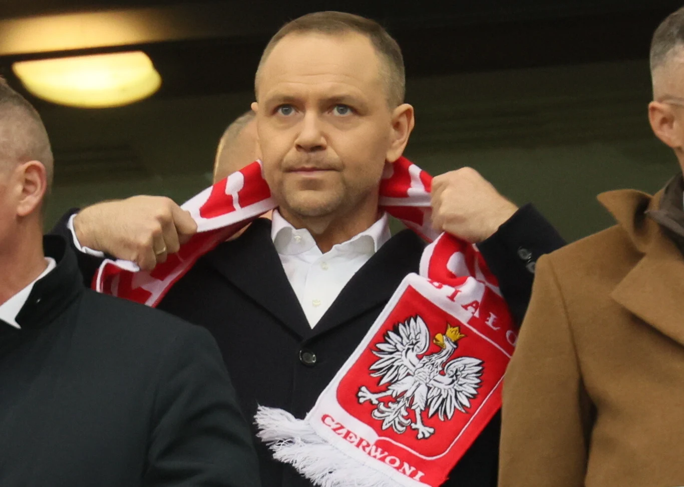 mężczyzna w eleganckim płaszczu trzyma na ramionach biało-czerwony szalik z godłem Polski i napisem 'Czerwoni', stojąc wśród innych osób na tle stadionu