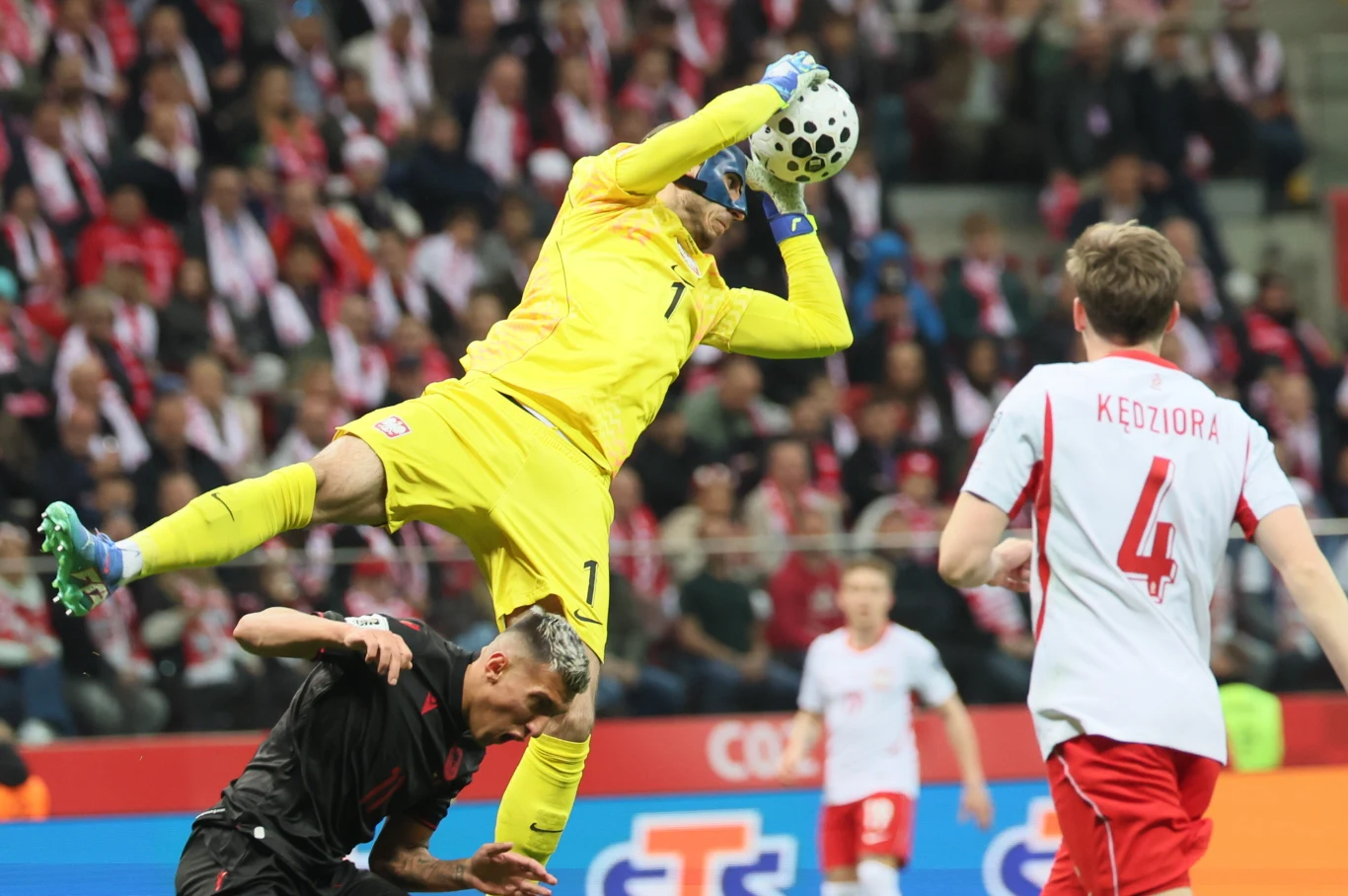 bramkarz w żółtym stroju łapie piłkę podczas meczu piłki nożnej, wyskakując nad zawodnikiem w czarnym stroju, obok piłkarz w białej koszulce z numerem 4 na plecach