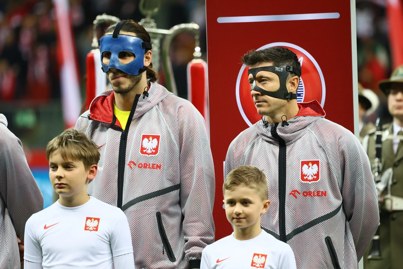 dwóch polskich piłkarzy w szaro-czerwonych reprezentacyjnych dresach z czarnymi i niebieskimi maskami ochronnymi na twarzach stoi obok siebie, przed nimi dwóch chłopców w białych koszulkach, w tle fragmenty stadionu i sylwetki żołnierzy