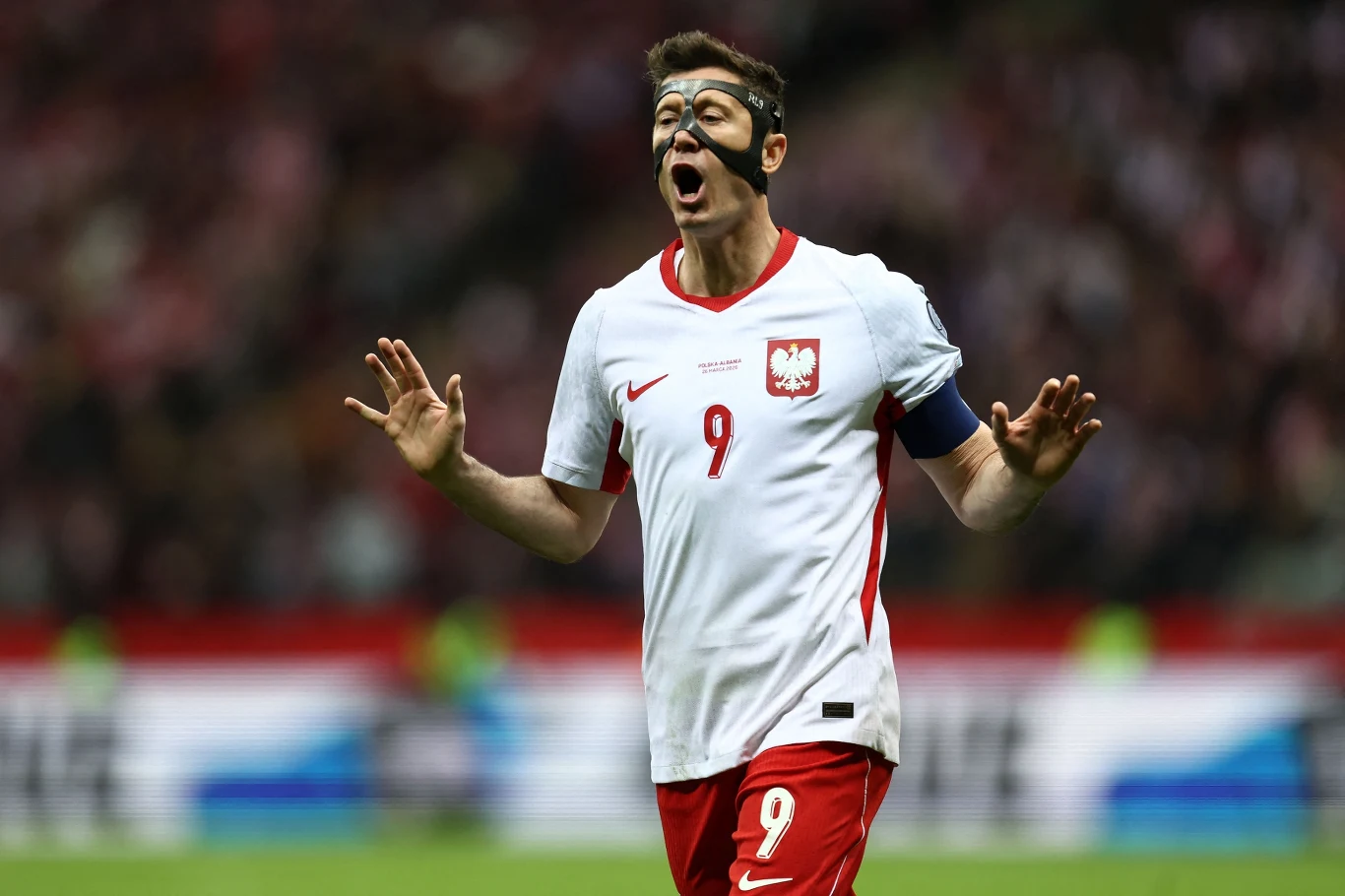 Piłkarz w białej koszulce polskiej reprezentacji z numerem 9 i opaską kapitana, noszący czarną maskę ochronną na twarzy, jest w trakcie dynamicznej reakcji na boisku, trzymając obie ręce uniesione w geście zatrzymania lub uspokojenia.