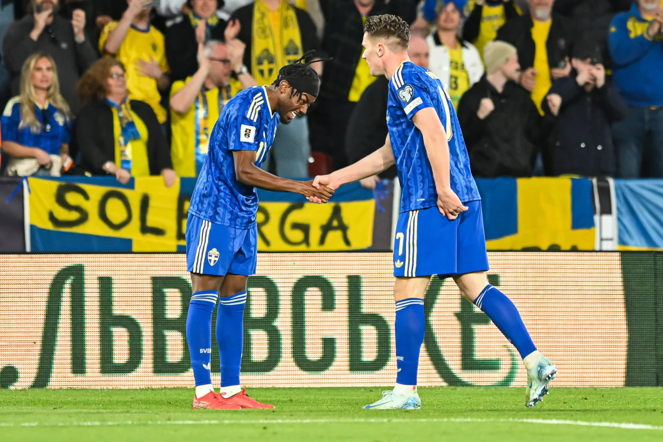 Dwóch piłkarzy w niebieskich strojach świętuje razem udaną akcję na murawie, wymieniając uścisk dłoni, podczas gdy w tle kibice ubrani w żółte barwy entuzjastycznie reagują na wydarzenia na boisku.