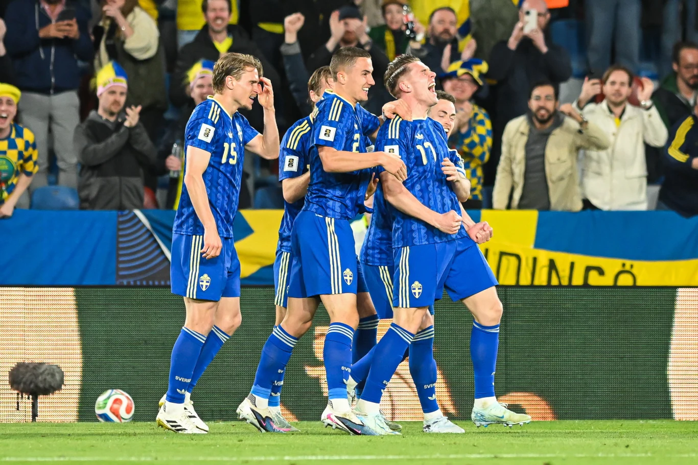 czterech piłkarzy w niebieskich strojach celebruje zdobycie gola na tle kibiców, którzy okazują radość i trzymają flagi w żółto-niebieskich barwach