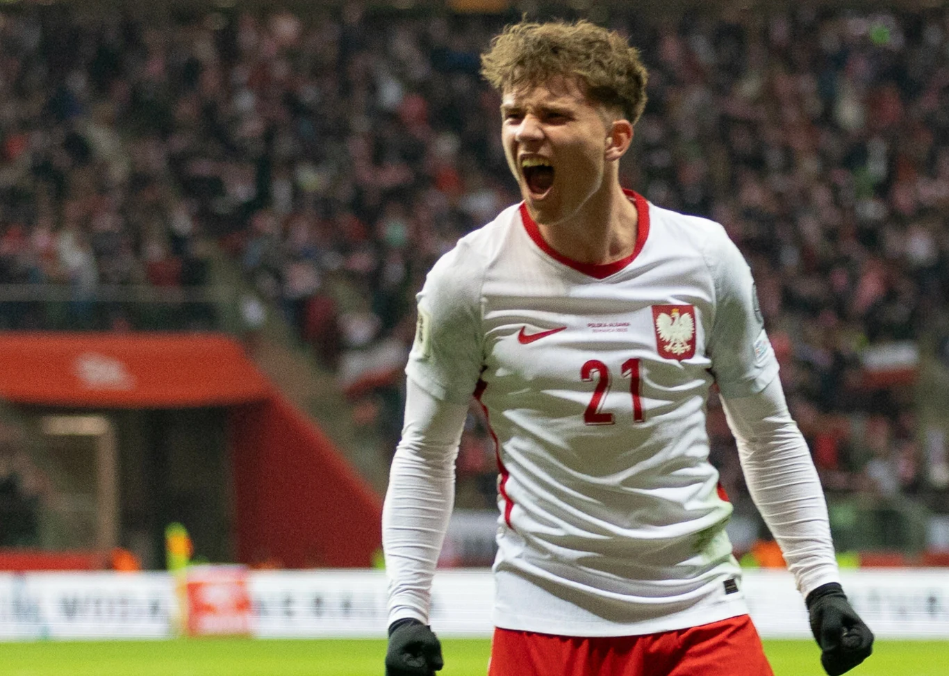 piłkarz w koszulce reprezentacji Polski z numerem 21 świętuje bramkę, wyrażając silne emocje na tle wypełnionego kibicami stadionu