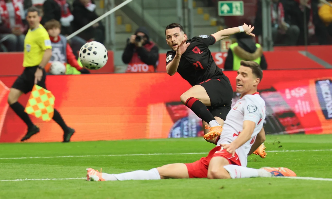 piłkarz w czarnej koszulce oddaje strzał na bramkę, podczas gdy zawodnik w białej koszulce wślizgiem próbuje temu zapobiec na tle stadionu wypełnionego kibicami