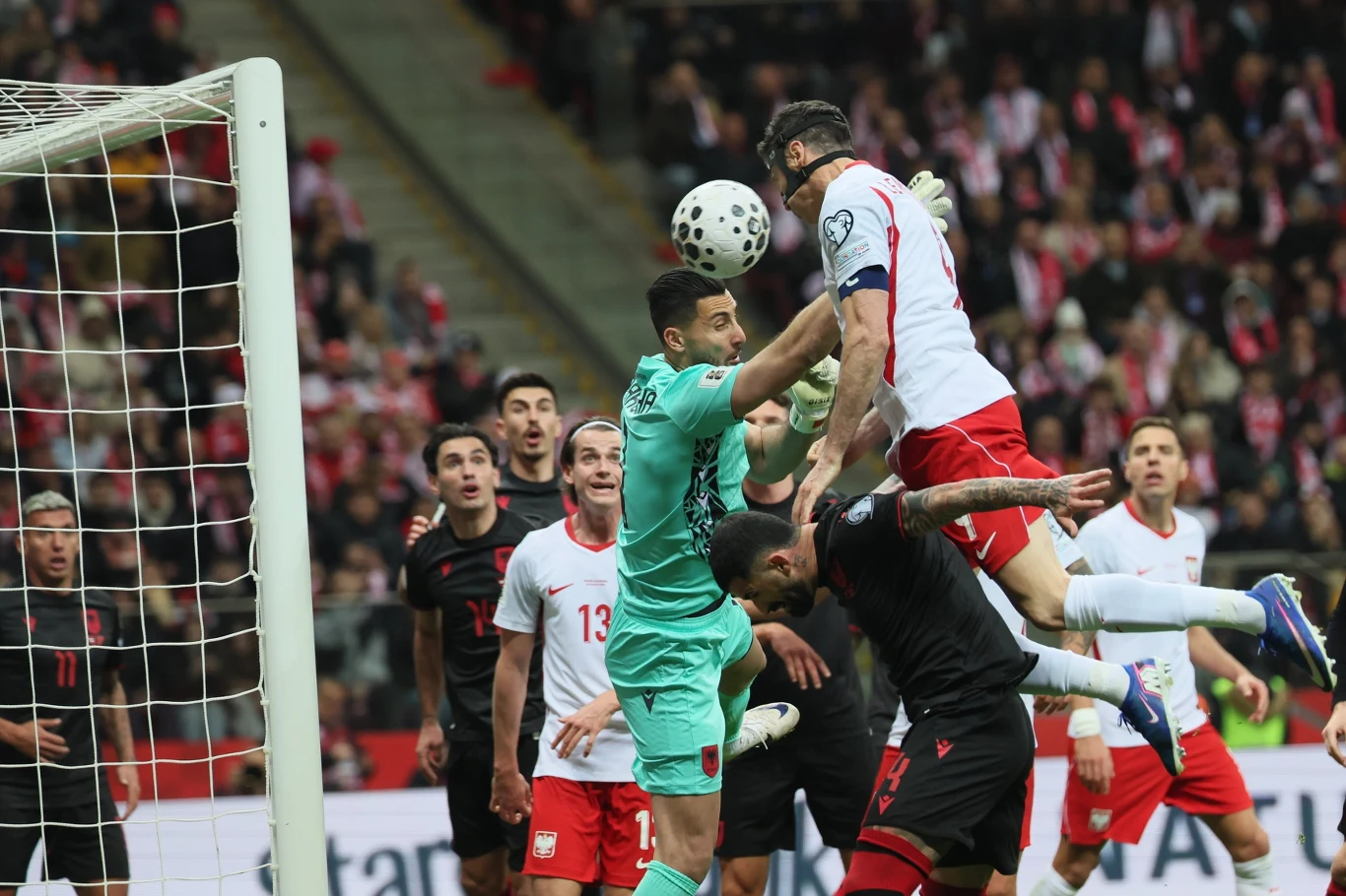 piłkarz w białoczerwonym stroju wyskakuje do główki podczas meczu piłki nożnej, w pobliżu bramki bramkarz w turkusowym stroju próbuje złapać piłkę, wokół zawodnicy obu drużyn obserwują sytuację