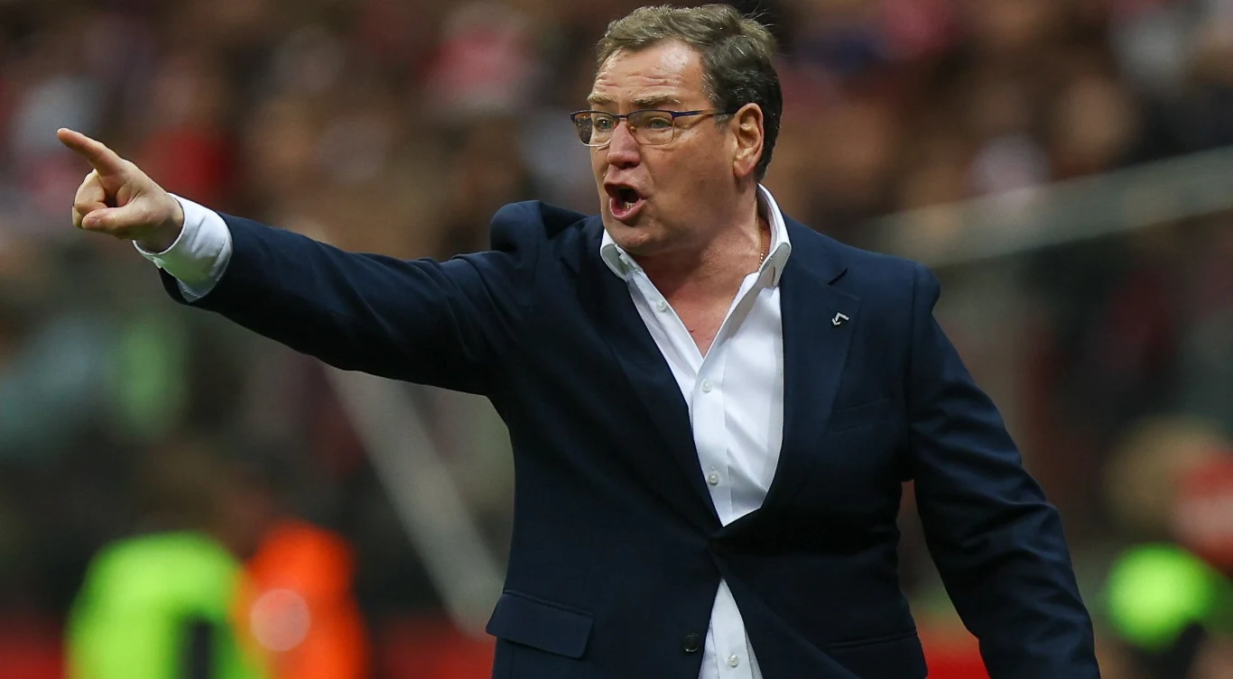 Mężczyzna w garniturze energicznie gestykuluje, wskazując wyciągniętą ręką w konkretnym kierunku, sugerując wydawanie poleceń lub motywowanie drużyny podczas wydarzenia sportowego.