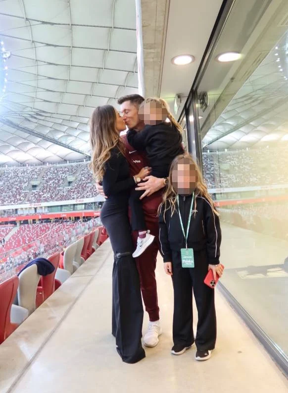 rodzina obejmująca się na tle stadionu sportowego, mężczyzna całuje kobietę, jedno dziecko jest trzymane na rękach, drugie stoi obok w sportowym stroju z akredytacją na szyi