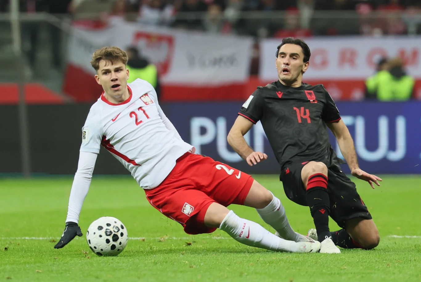 dwóch piłkarzy walczy o piłkę podczas meczu, zawodnik w białym stroju z numerem 21 wykonuje wślizg, a zawodnik w czarnym stroju z numerem 14 stara się przejąć piłkę