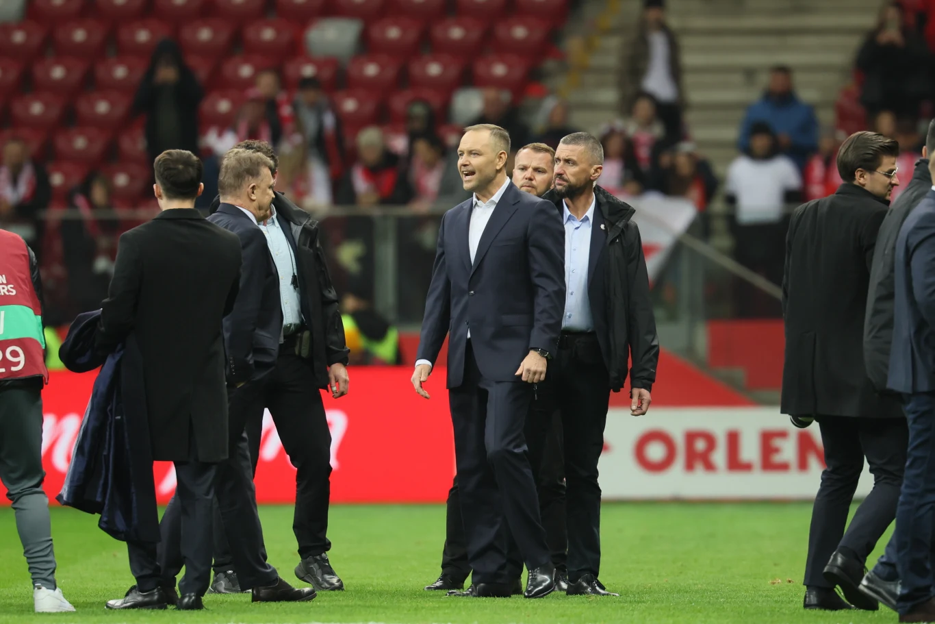 grupa mężczyzn w garniturach przechodzi przez murawę stadionu, w tle widoczni kibice na trybunach oraz wyraźny baner reklamowy