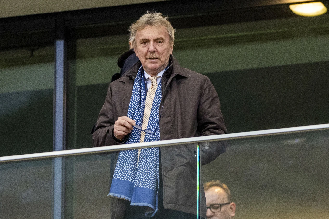 starszy mężczyzna w płaszczu i niebieskim szaliku stoi na trybunach, trzymając okulary w dłoni, za szklaną barierką