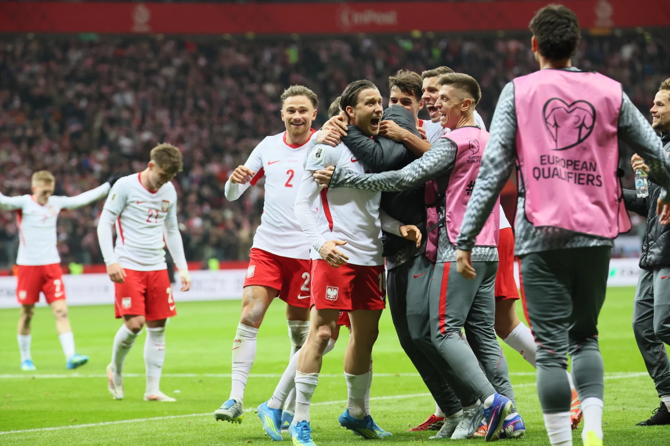 grupa polskich piłkarzy w biało-czerwonych strojach świętuje zdobycie gola na stadionie, piłkarze są wyraźnie podekscytowani i otoczeni przez kolegów z drużyny, w tle widać tłum kibiców na trybunach