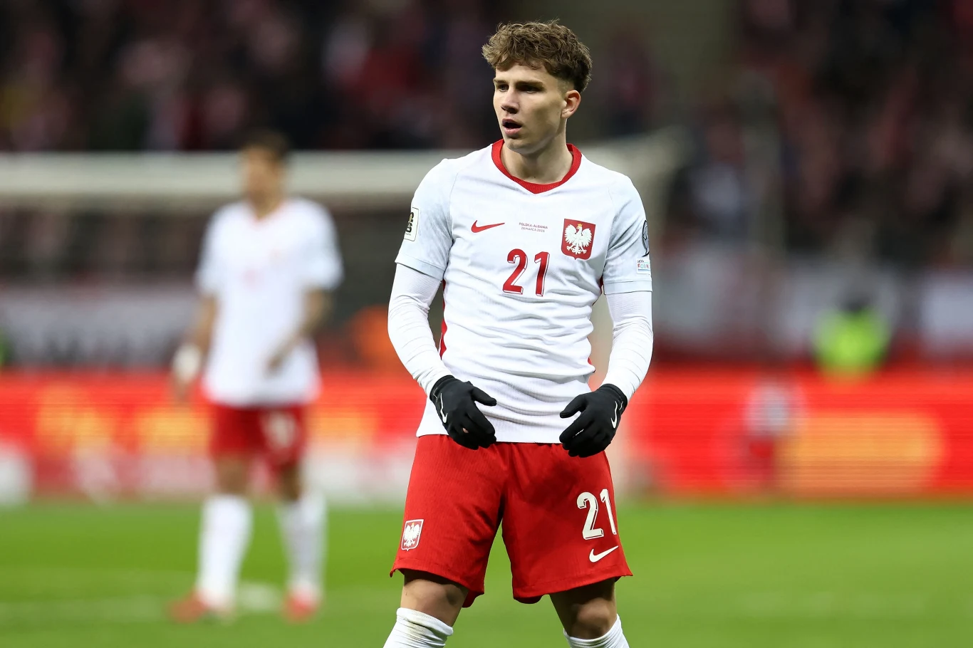 polski piłkarz w białej koszulce z numerem 21 i godłem narodowym, skupiony podczas meczu na stadionie, w tle rozmyte postaci innych zawodników oraz trybuny z kibicami