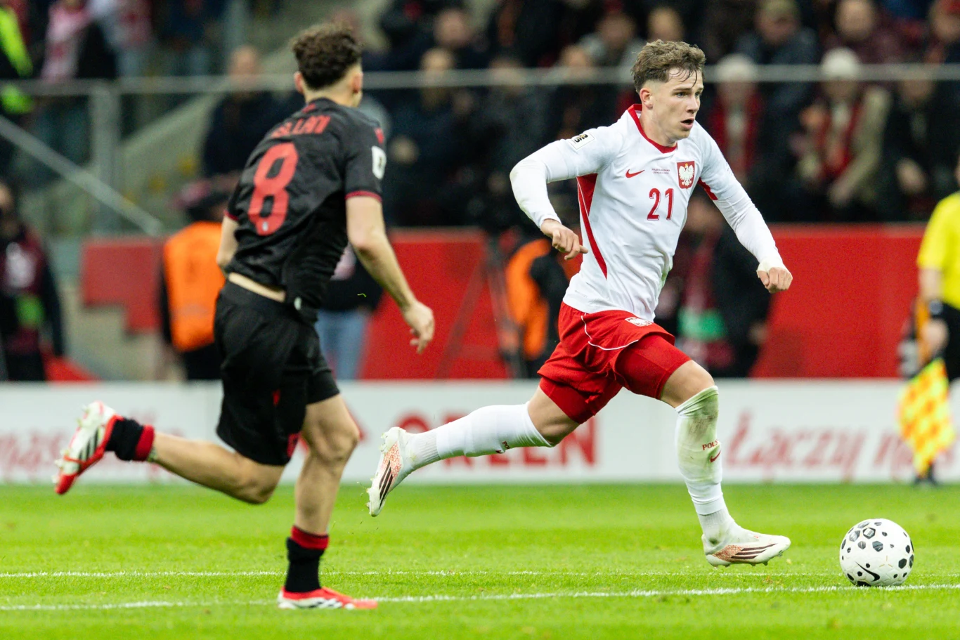 piłkarz w biało-czerwonym stroju reprezentacji Polski dynamicznie biegnie z piłką obok zawodnika drużyny przeciwnej podczas meczu na stadionie, na tle widocznych trybun z kibicami