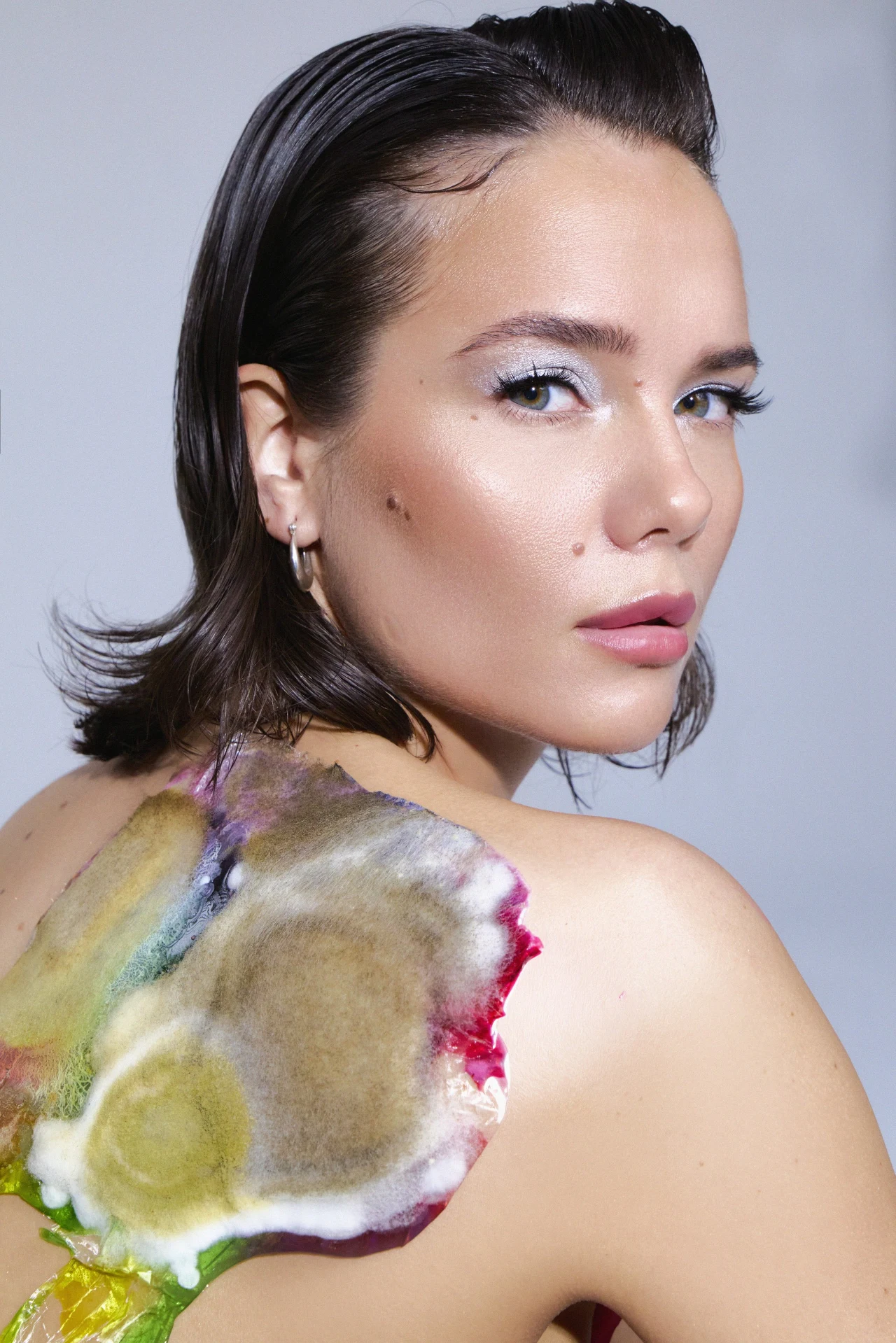 kobieta z krótkimi, ciemnobrązowymi włosami, spojrzeniem skierowanym przez ramię, z delikatnym makijażem i wyrazistym, kolorowym wzorem na ramieniu przypominającym plamy farby lub abstrakcyjną sztukę