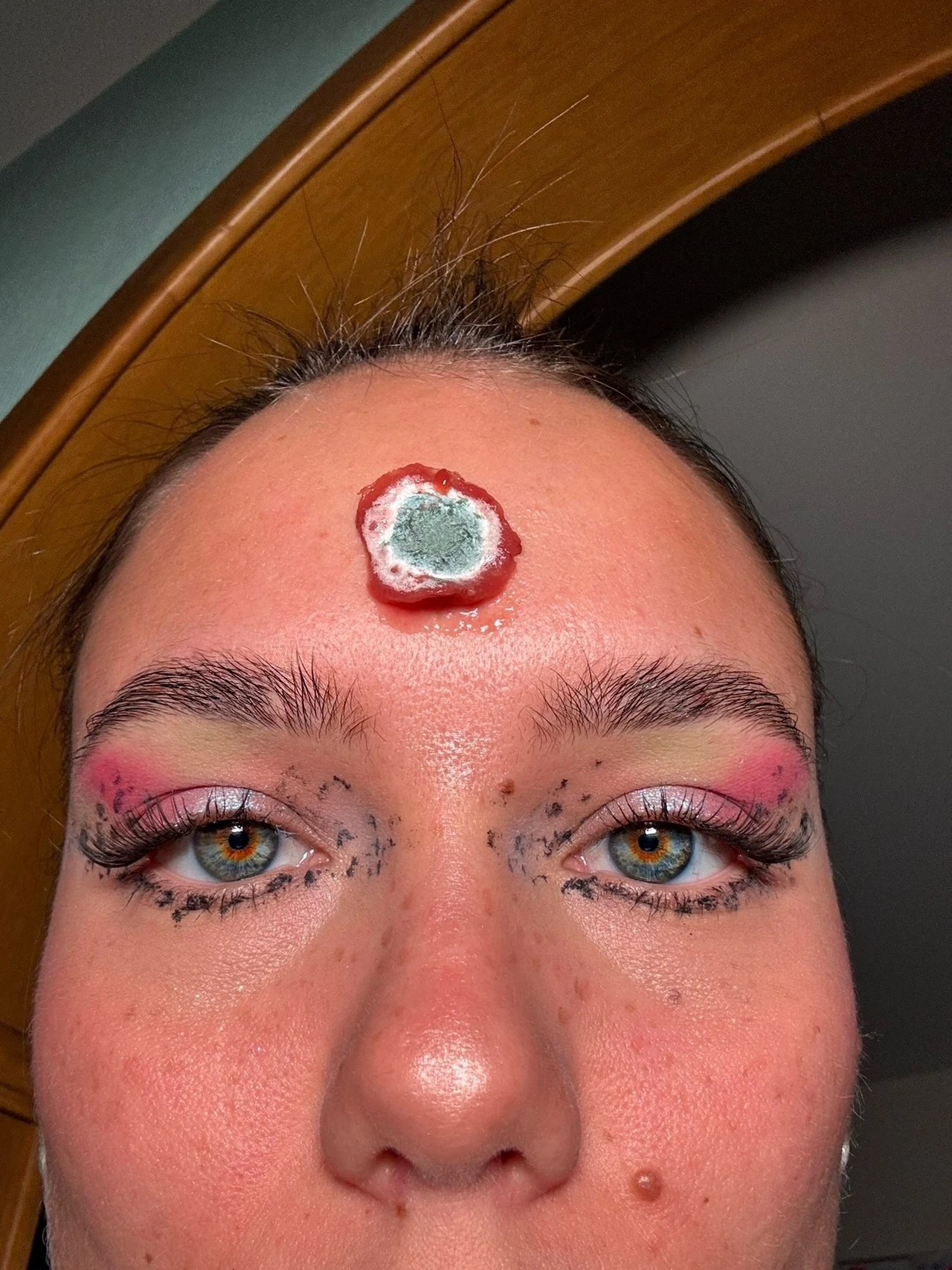 kobieta z mocnym makijażem oczu oraz efektowną, sztuczną raną na środku czoła stylizowaną na wrzód lub ranę z pleśnią, co sugeruje inspirację charakteryzacją filmową lub halloweenową