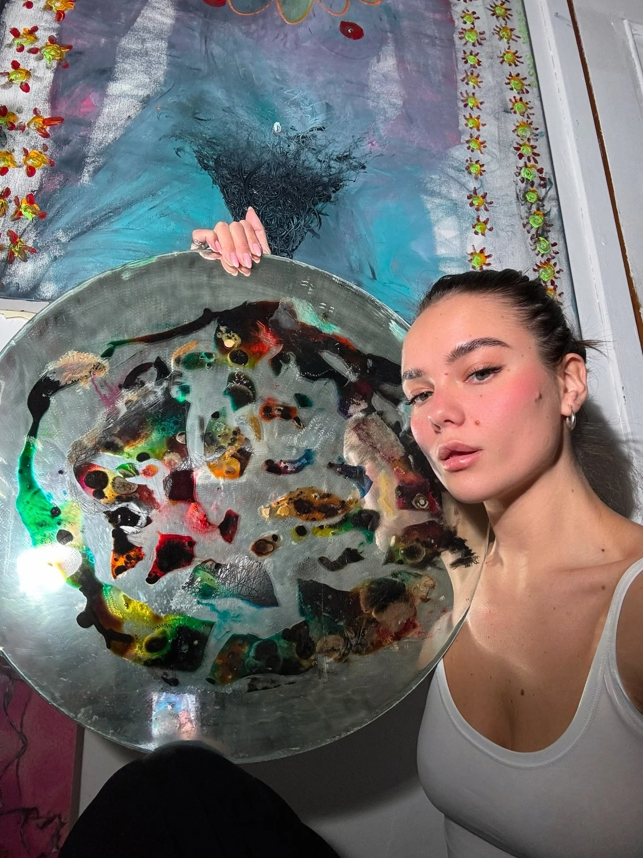 Młoda kobieta w białym topie pozuje z dużym okrągłym dziełem sztuki z abstrakcyjnymi, kolorowymi wzorami, w tle widoczny fragment obrazu z motywem niebieskiego rozprysku.