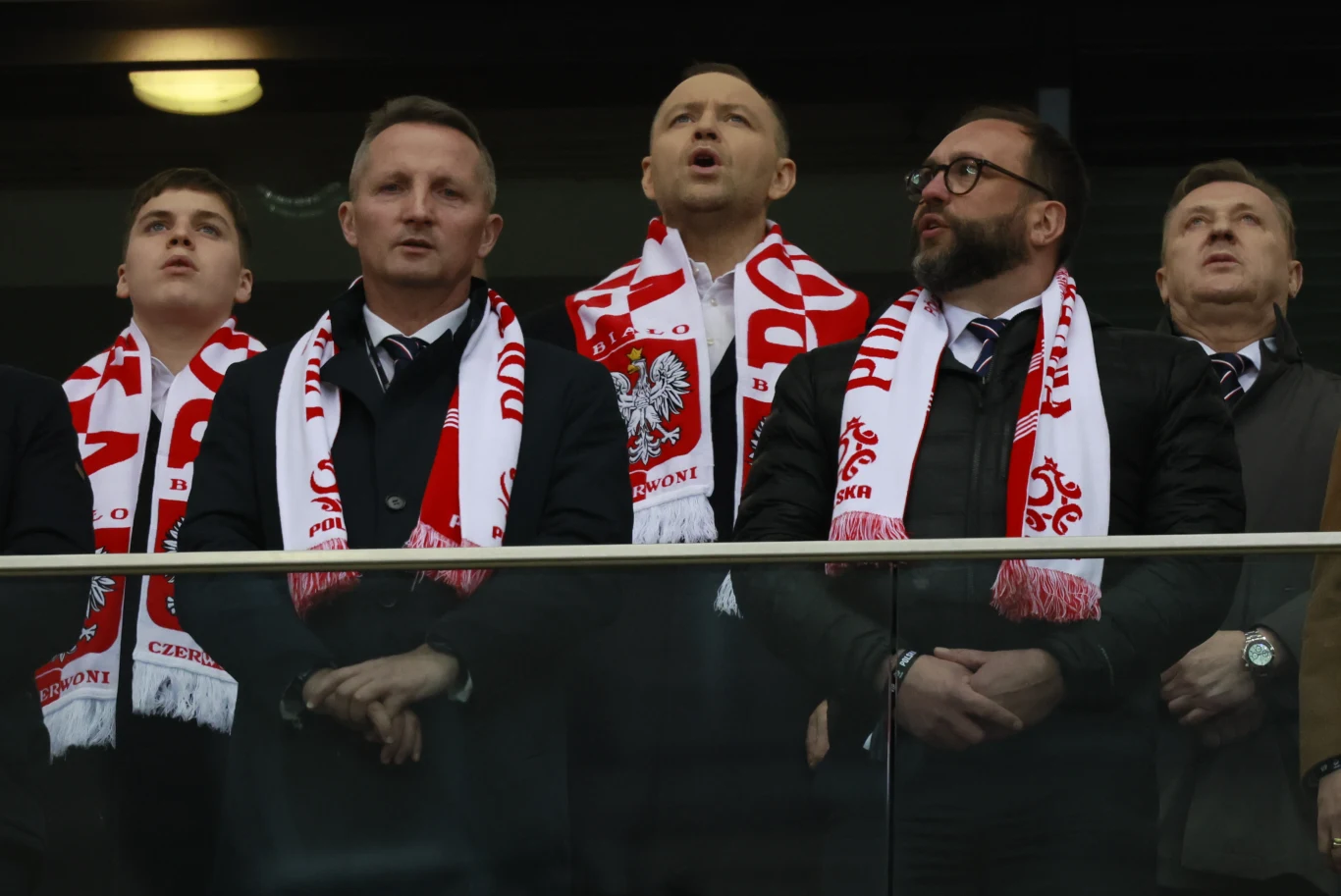 Grupa mężczyzn ubranych w garnitury i biało-czerwone szaliki z godłem Polski oraz napisami kibicowskimi śpiewa hymn lub doping na stadionie, stojąc za szklaną barierką.
