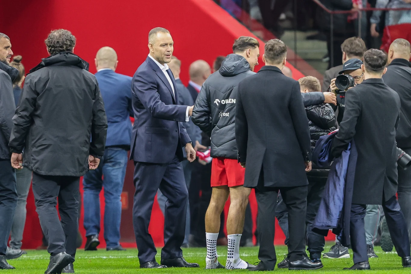 Grupa mężczyzn w eleganckich garniturach oraz stroju sportowym stoi na murawie stadionu, w centrum uwagi jest zawodnik w czerwonych spodenkach i sportowej kurtce otoczony przez rozmówców i kamerzystę, dominują żywe barwy czerwieni oraz atmosfera rozmów...