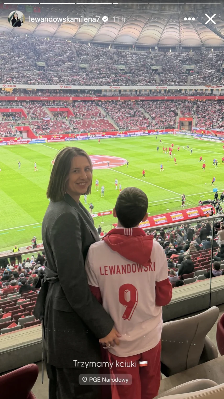 kobieta obejmująca dziecko w koszulce piłkarskiej z nazwiskiem Lewandowski na tle pełnego stadionu podczas meczu piłkarskiego, tłum kibiców skoncentrowany na wydarzeniach na murawie