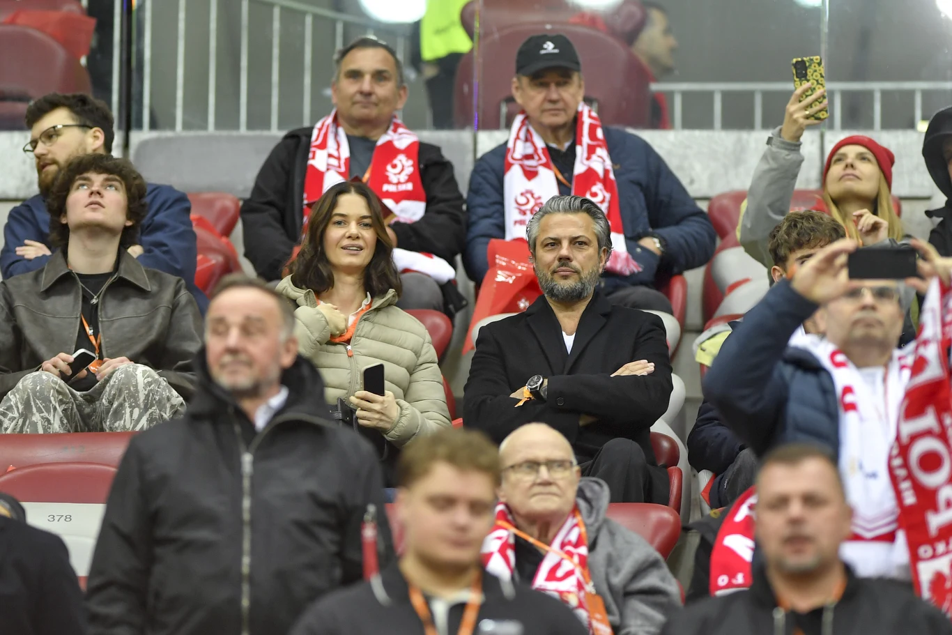 Grupa kibiców w czerwono-białych szalikach siedzi na trybunach stadionu podczas meczu piłkarskiego; niektórzy oglądają wydarzenia na boisku, inni robią zdjęcia lub rozmawiają, a w centrum kadru znajduje się mężczyzna z siwymi włosami i brodą, z założon...