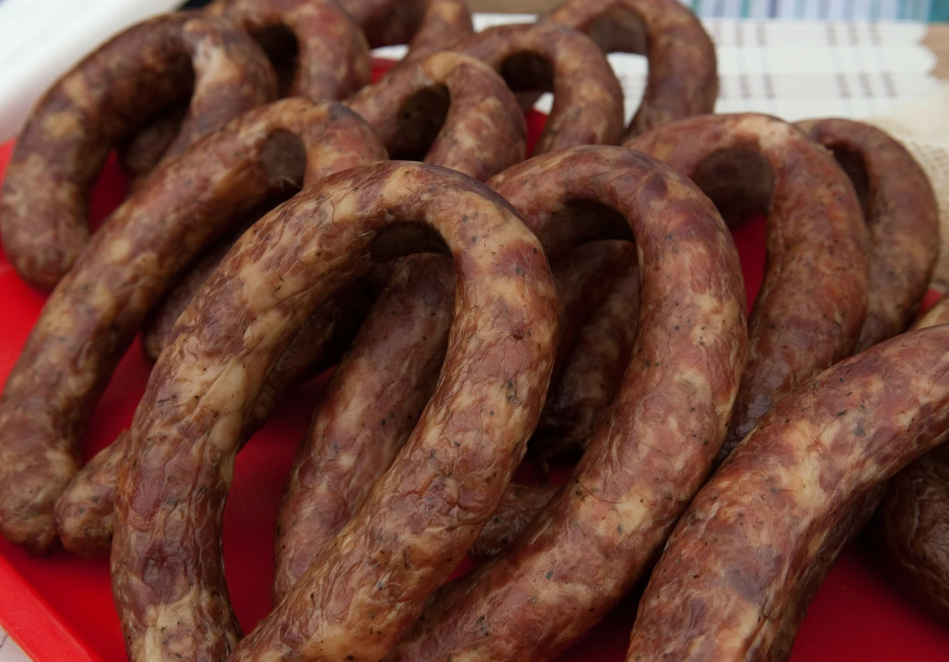 Jak przechowywać kiełbasę? Tak lodówka "nie przejdzie" niepożądanym zapachem. Kilka pęt kiełbasy o ciemnej barwie na czerwonej tacy, prezentujące sposoby przechowywania w lodówce.