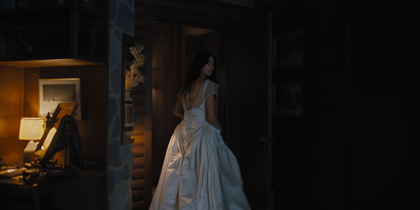 kobieta w eleganckiej, białej sukni z odkrytymi plecami stoi w półmroku przy uchylonych drzwiach, delikatnie odwracając się w stronę światła bijącego z przytulnego kącika z lampką, zdjęciem i stojącym na biurku wazonem
