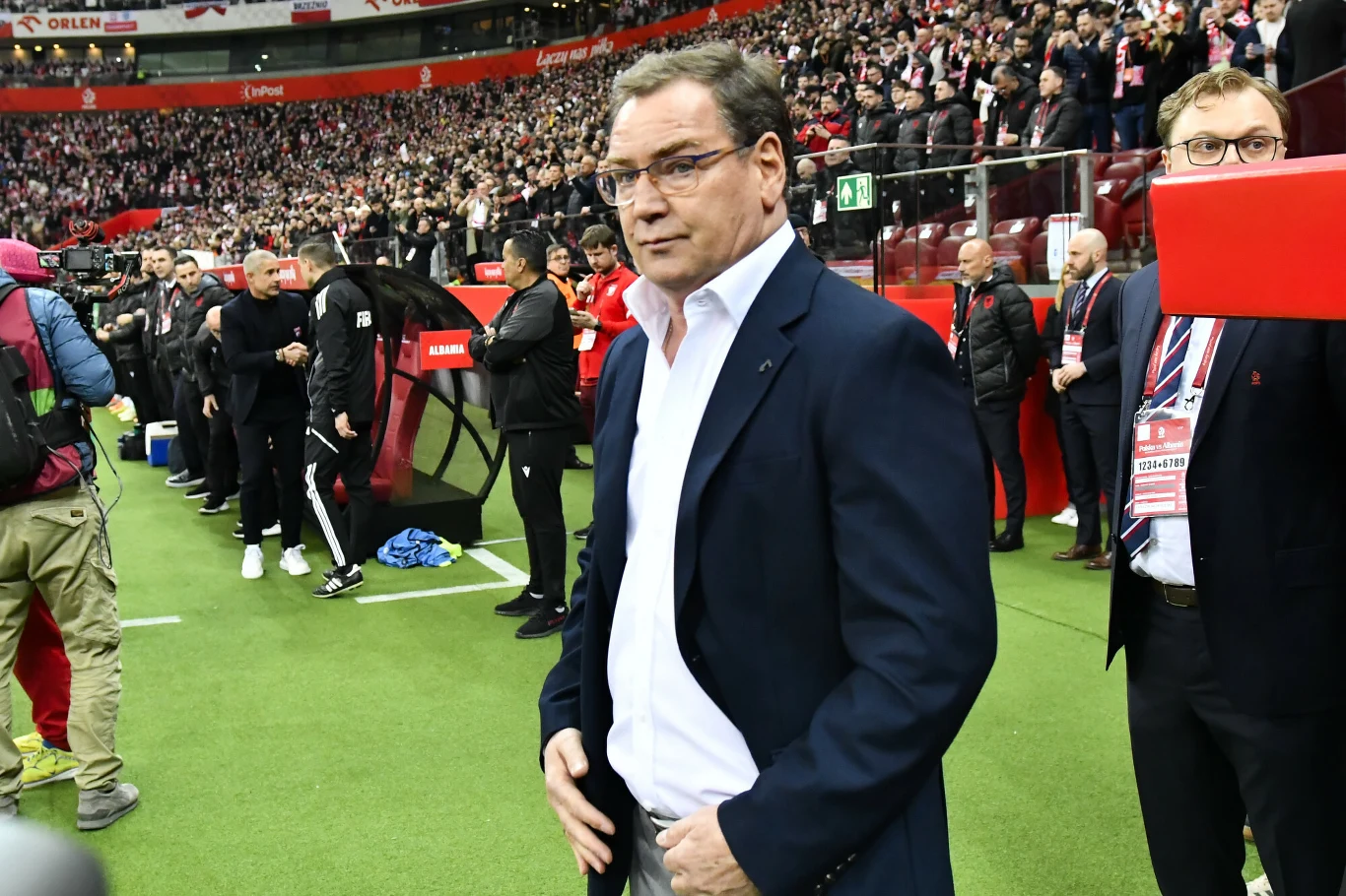 mężczyzna w garniturze stoi na zielonej murawie stadionu piłkarskiego, wokół niego znajduje się wielu ludzi, a w tle widoczny jest tłum kibiców na trybunach