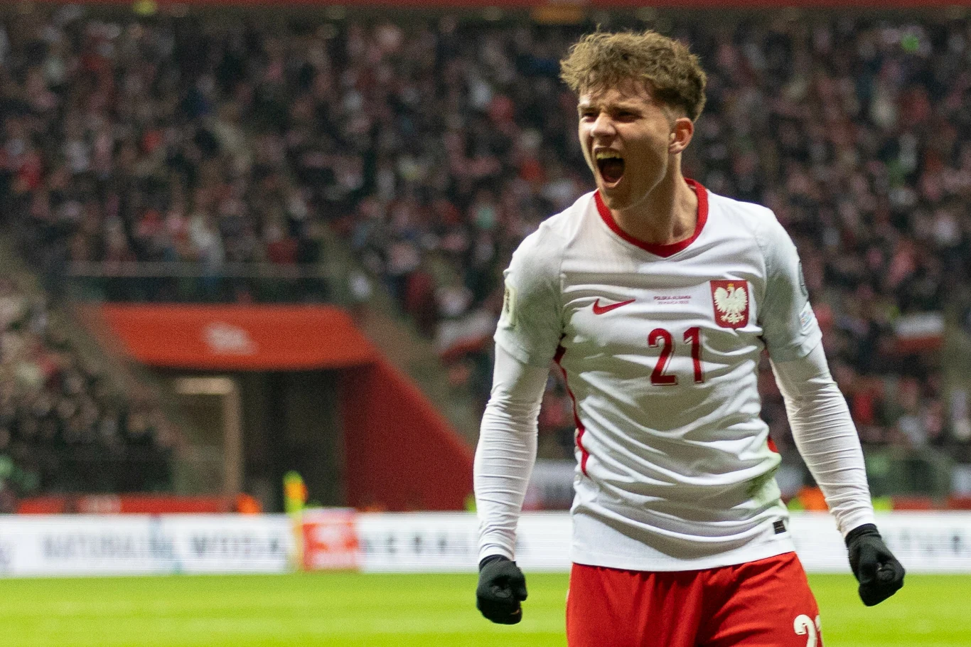 piłkarz w koszulce reprezentacji Polski z numerem 21 świętuje zdobycie gola na tle rozemocjonowanych kibiców na stadionie