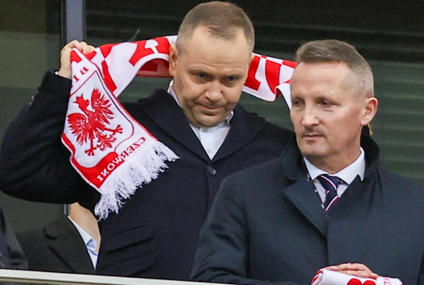 Dwóch mężczyzn ubranych elegancko na trybunach stadionu, jeden trzyma szalik z godłem Polski i napisem 'POLSKA'.