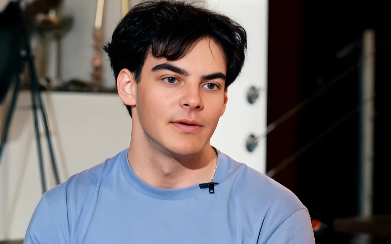 Kacper Walas, przewodnik Michała Gołasia Młody mężczyzna z ciemnymi włosami i gładką cerą w niebieskiej koszulce patrzy lekko w bok, ubrany w przypięty mikrofon, w tle niewyraźne elementy wyposażenia wnętrza.