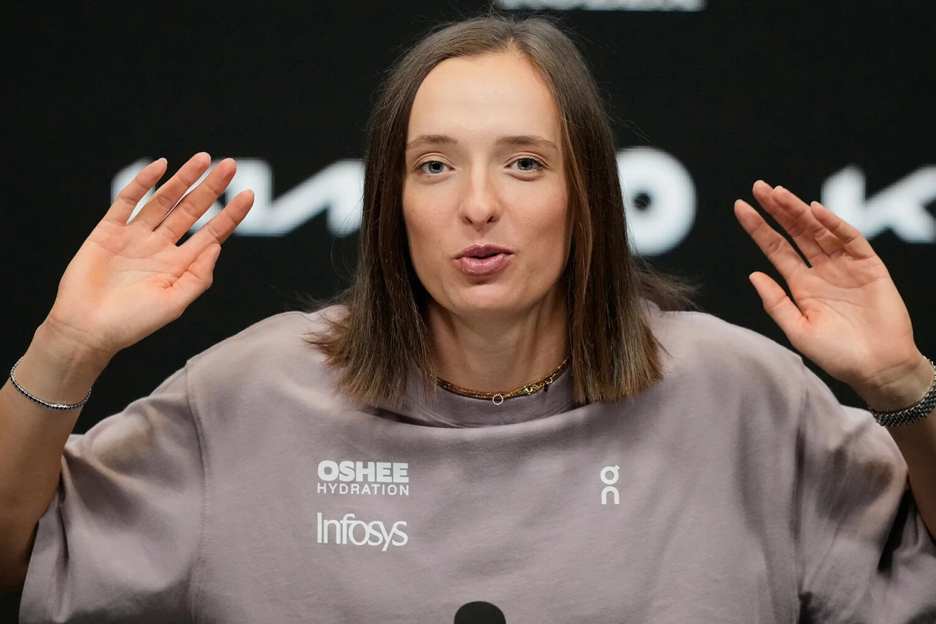 kobieta z prostymi, brązowymi włosami w jasnej bluzie z logotypami, trzyma uniesione dłonie na wysokości głowy, siedzi przed mikrofonem na tle ciemnej ściany z białymi napisami