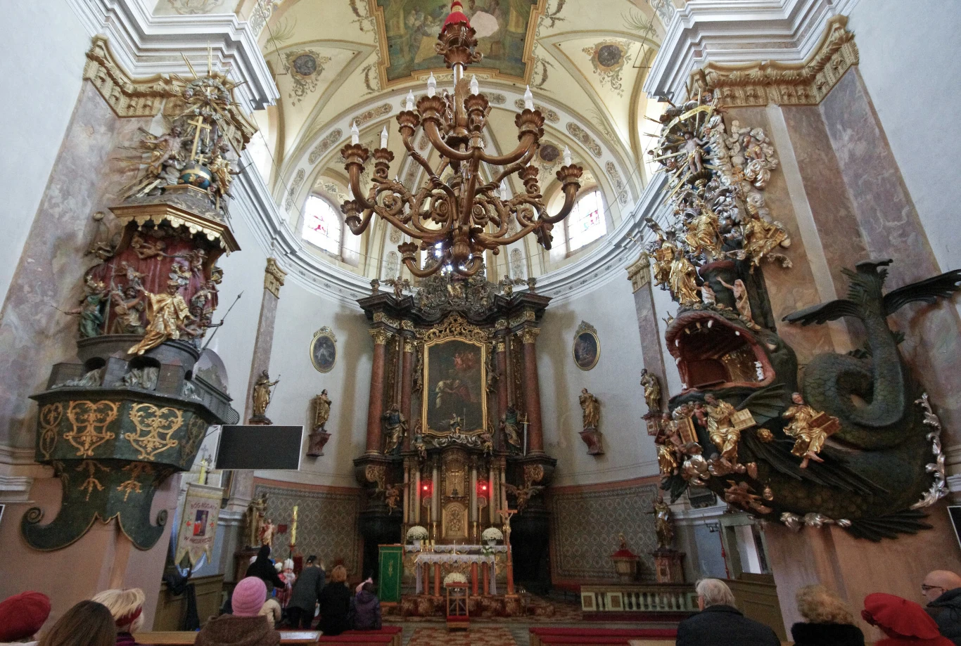 bogato zdobione wnętrze kościoła z dużym żyrandolem pośrodku, barokowym ołtarzem frontalnym oraz dwiema imponującymi ambonami – jedna w formie statku, druga przedstawiająca smoka z otwartą paszczą; przed ołtarzem zgromadzeni wierni
