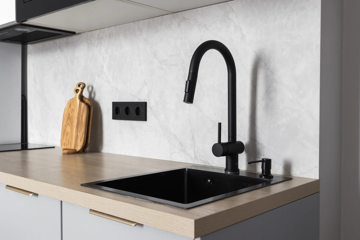 Imitacja betonu to jeden z najpopularniejszych wzorów tapet do kuchni. nowoczesny blat kuchenny z czarnym zlewem i baterią, na blacie drewniana deska do krojenia, w tle marmurowa ściana z trzema czarnymi gniazdkami elektrycznymi