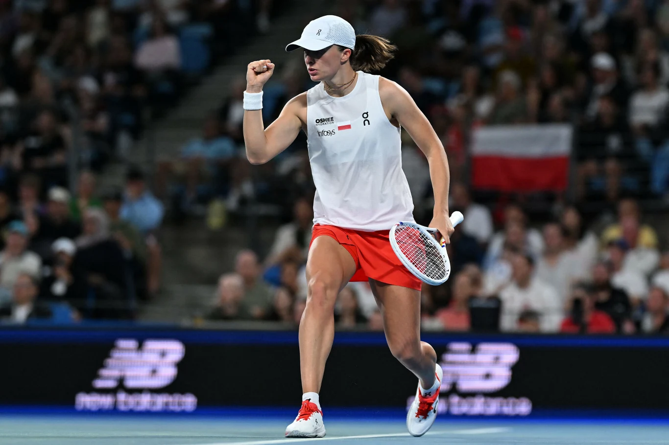 Tenisistka celebrująca zdobyty punkt na korcie, ubrana w biały top, czerwone spodenki i białą czapkę z daszkiem, w tle widoczna polska flaga oraz publiczność na trybunach.