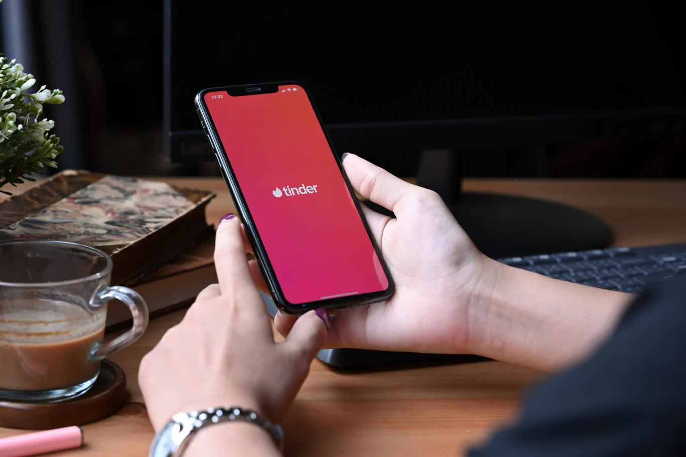 osoba trzymająca w dłoni smartfon z uruchomioną aplikacją Tinder, obok na biurku filiżanka kawy, książka i komputer