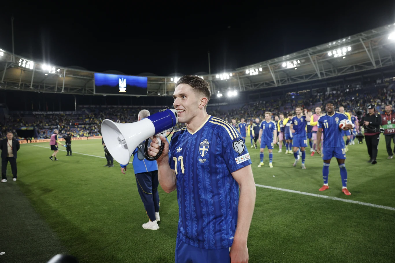 piłkarz w niebieskiej koszulce z numerem 11 trzyma megafon i przemawia do kibiców na stadionie, wokół niego znajdują się inni zawodnicy w podobnych strojach, a w tle widać trybuny z publicznością oraz oświetlony stadion