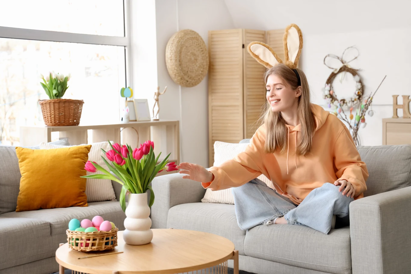 Niektóre przesądy wielkanocne wydają się dziś dziwne, ale dawniej ściśle ich przestrzegano. Sprawdź, czego nie robić w Wielką Niedzielę i co robić w święta, by zapewnić sobie szczęście. Młoda kobieta w opasce z króliczymi uszami siedzi na kanapie i sięga ręką do bukietu tulipanów. Wielkanocny wystrój w tle.