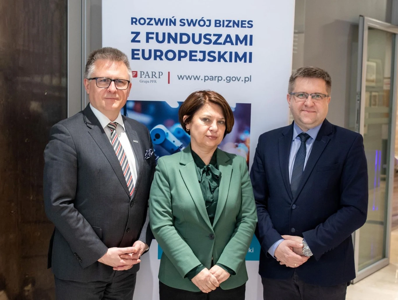Trzy osoby w eleganckich strojach stoją obok siebie na tle roll-upu promującego rozwój biznesu z funduszami europejskimi, pośrodku kobieta w zielonej marynarce, po bokach dwóch mężczyzn w garniturach.