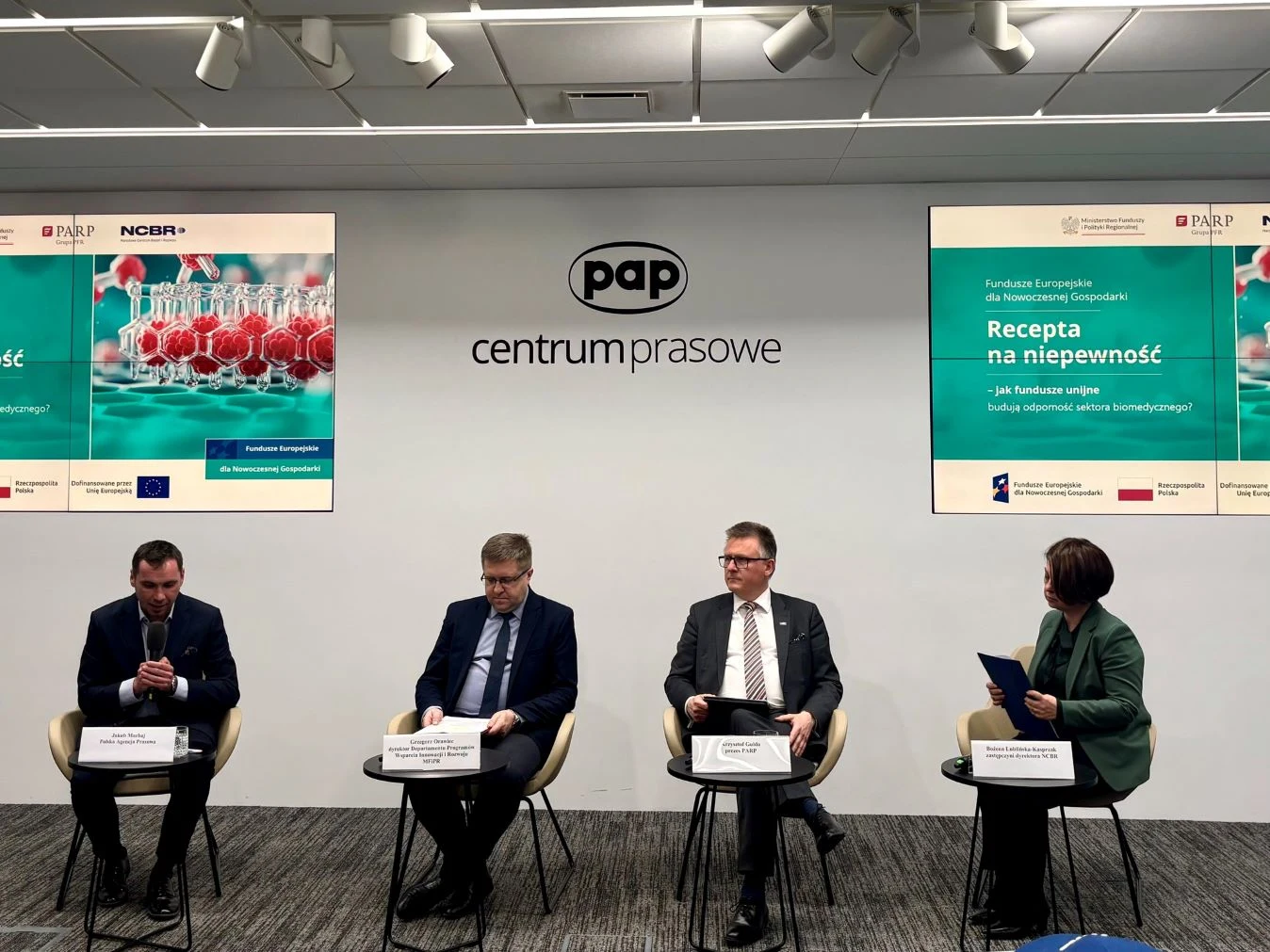 czterech uczestników w garniturach i jednej kobiety siedzi na scenie podczas panelu dyskusyjnego w centrum prasowym PAP, za nimi na ścianie umieszczone są dwa duże ekrany prezentujące temat spotkania 'Recepta na niepewność' oraz logotypy organizatorów