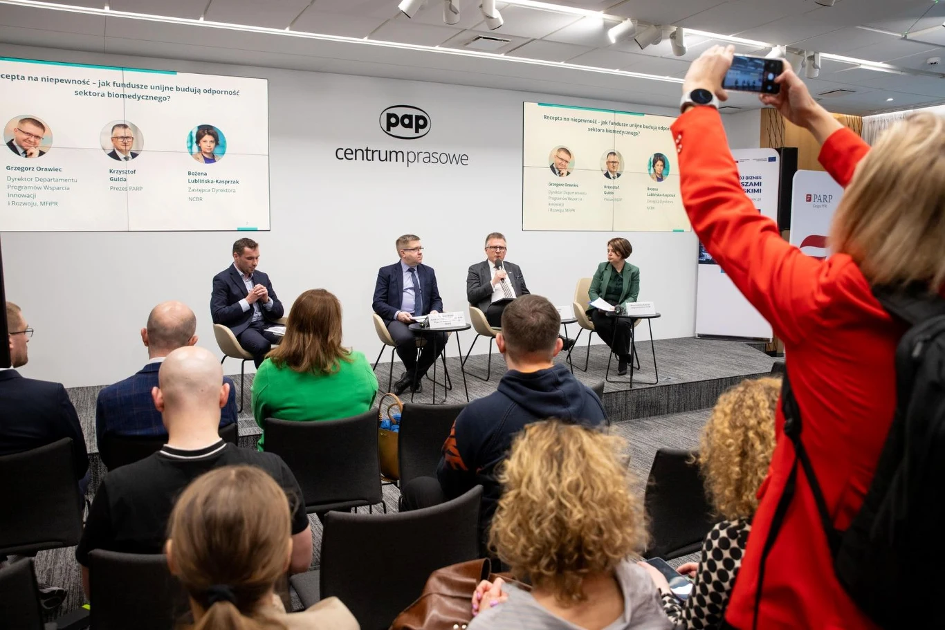 Panel ekspertów siedzi na scenie w centrum prasowym, omawiając temat widoczny na prezentacji z tyłu, podczas gdy publiczność słucha i jedna osoba fotografuje wydarzenie smartfonem.