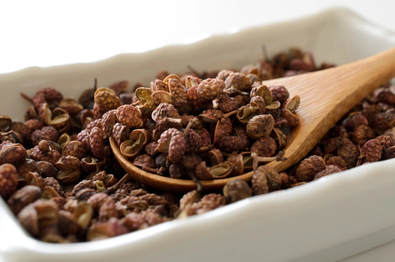 Pieprz syczuański świetnie podkreśla smak mięs Drewniana łyżka na białym naczyniu z suszonym pieprzem syczuańskim o brązowo-czerwonym zabarwieniu.