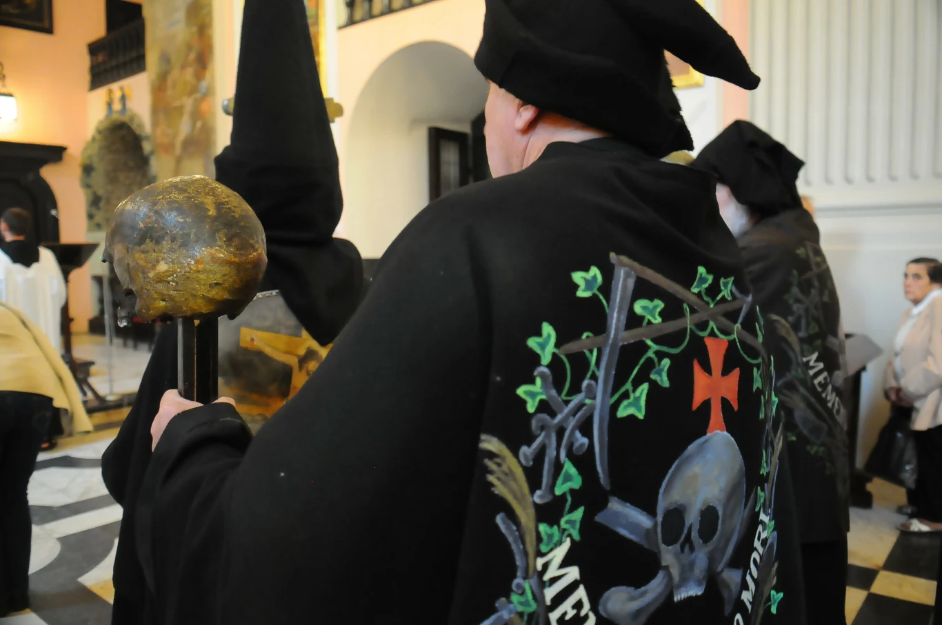 Postać w czarnym kapturze z wyhaftowanym symbolem czaszki i mieczem na płaszczu, trzymająca ozdobiony kij, otoczona innymi osobami w podobnych strojach, w tle widoczne wnętrze budynku z łukami i kolumnami.