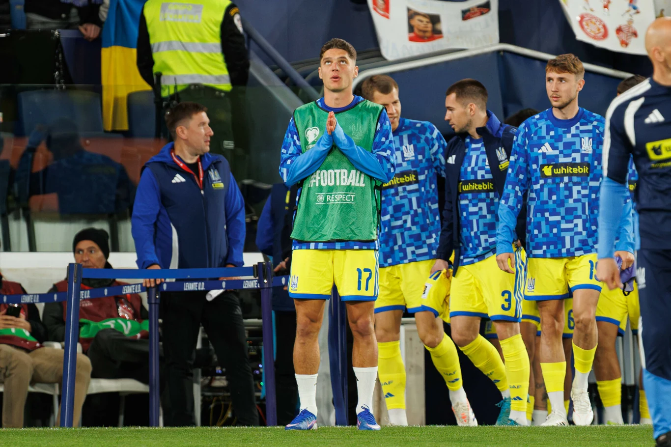 piłkarze w żółto-niebieskich strojach idą razem po murawie, jeden z nich stoi na pierwszym planie z założonymi rękami, w tle widoczni trenerzy oraz fragment flag i stadionu